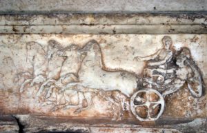 Ancient Roman marble relief sculpture depicting chariot race scene with horses and driver carved in deteriorated stone panel