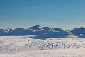 Cold desert landscape with snow-covered terrain and ice ridges stretching toward distant mountains under a clear blue sky.