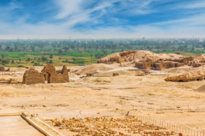 Desert archaeological landscape with ancient ruins and sandy hills in the foreground, overlooking a green river valley and distant city skyline under a blue sky.